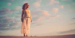 beautiful aging woman with curly brown hair stands on sand in front of vibrant sky