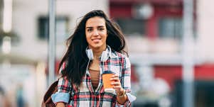Young Latina woman crossing a city street with coffee and headphones