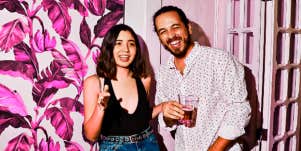 young couple at party against pink floral background, laughing, her hand on his arm