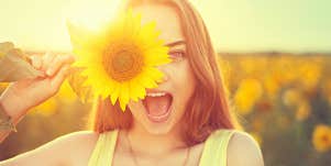 Young woman in a sunflower field smiling behind a flower, sunlit from behind