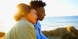 lovely couple with brown skin stares out at the ocean, backlit 