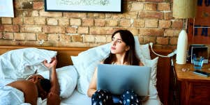 woman looking worried and holding her laptop in bed while man is asleep next to her