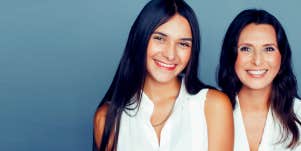 Brunette mother and adult daughter with olive skin tone and white collar shirts smile at camera