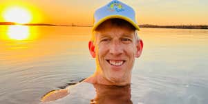 58 year old white man in ball cap in still water, sunset behind him