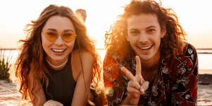 young couple smiles at camera lying on the beach, man giving peace sign
