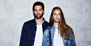 couple standing against a grey wall, both wearing denim jackets and white tees