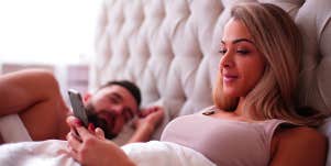 woman laying in bed looking at phone with man sleeping next to her