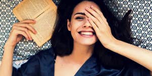 brunette woman smiling, hand over one eye, book in hand on a bed