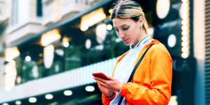 young blonde woman works on phone in busy street in orange windbreaker