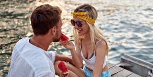 Young blonde couple on a dock, happily feeding each other watermelon