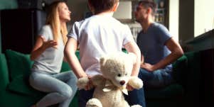 parents sitting on couch fighting in front of little boy holding a teddy bear