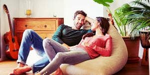 couple lounges in the sunlight, resting on a large pouf chair, looking at one another