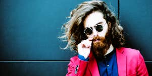 man with long hair wearing bright pink suit plays with his hipster mustache