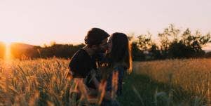 man and woman, each with brown hair, kiss in a field at sunset