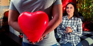 man approaching woman while holding big red plastic heart between his hands behind his back to present to her