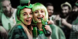 two young white women in St. Patrick's Day garb, hold beers in a bar