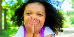 young girl with brown skin and black hair covers her mouth and grins