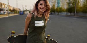 Cheerful girl with tattoos posing with a longboard while walking on the road in the city