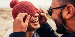 Loving couple having fun outdoors. Man covering eyes of woman with cap.