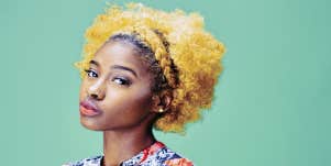 Black woman with blonde hair against green background, looking at camera