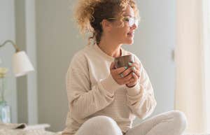 Beautiful caucasian young woman with curly hair and eyeglasses sitting on bed and looking away while drinking coffee in morning
