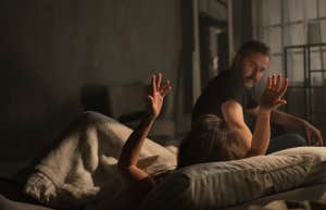 boyfriend sitting on bed introgating girlfriend, who throws hands in air