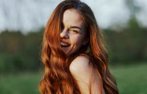 A young woman smiles and looks into the camera with her long, red, wavy, shiny hair in a park with green grass