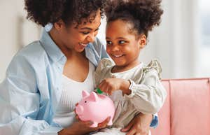 mom holding daughter in her lap putting money in piggy bank