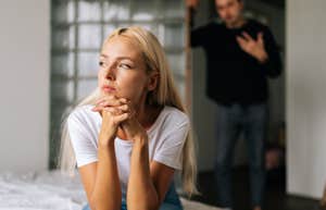 upset woman sitting on bed while man stands behind her trying to console her