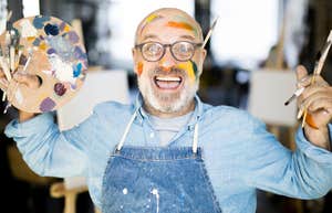 Ecstatic mature artist holding a palette and paintbrushes showing the art of starting over and the wild choices that completely rewrite your life.
