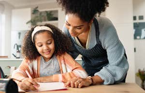 A mother helps her young daughter write in a notebook at a kitchen table.
