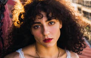 A close-up portrait of a woman with curly hair looking at the camera outdoors.