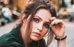A close-up portrait of a young woman with tousled hair resting her hand on her forehead outdoors.