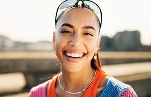 A woman smiling outdoors in sunlight with buildings in the background.