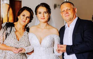 Happy parents hold the hands of the bride who is getting married and support her, wearing a wedding dress and a long veil.