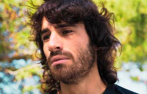 A somber man with long hair sitting alone in nature, appearing depressed and overwhelmed; illustrating the 'unseen' emotional struggle of men facing social disconnection in a modern landscape.