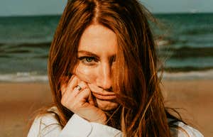 A woman with a sad expression stands on a scenic beach, illustrating the internal disconnect of feeling lonely even when life appears full on the surface.