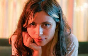 A woman sitting on a couch leaning forward with her hand on her chin, holding direct eye contact with a slightly sinister grin.
