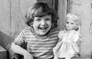 A retro black-and-white style snapshot of a young child playing alone with a toy doll near a balcony railing; illustrating 'the architectural change in play' where historic youth independence contrasts with modern supervised childhoods.