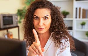 A close-up portrait of a corporate employee giving a firm warning gesture from her modern desk with plants and shelves behind her; illustrating 'the intellectual guardrail' model where setting limits signals high cognitive capability.