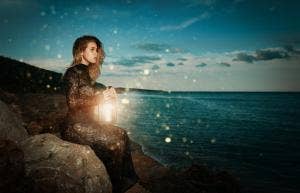 witchy looking woman sits on a rock. at sunset, looking at ocean